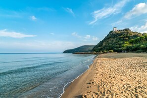 A beach with clear water, a sandy shore, and a building on a hill. - Villetta Il Pino (Castiglione della Pescaia)