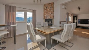 Un salon moderne avec une table à manger au plateau en verre, des fauteuils en cuir blanc, une cheminée en pierre, un téléviseur et un balcon agrémenté de rideaux.