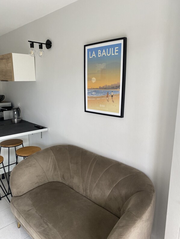 Un salon moderne avec un canapé beige, un coin cuisine équipé d’une machine à café et une affiche encadrée représentant « LA BAULE » accrochée au mur.