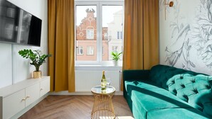 A modern living room with a teal sofa, a TV mounted on the wall, a small round table with a bottle and glass, and a window with curtains. - Elite Apartments Ogarna Prestige (Gdansk)