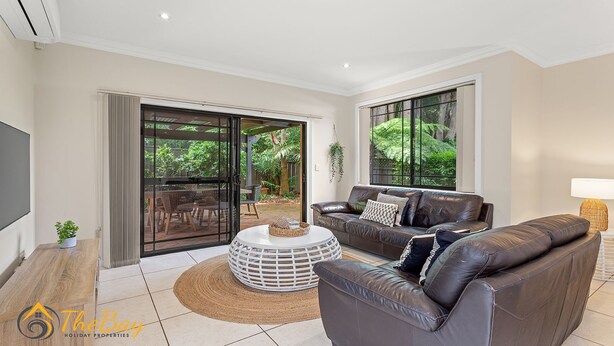 A modern living room with a leather sofa, a coffee table, and a TV. There are sliding glass doors leading to an outdoor patio with a dining area.