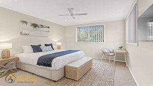 A bedroom with a bed, bedside tables, a desk, a chair, a ceiling fan, a window with blinds, and a beach-themed painting on the wall.