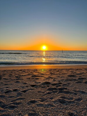 A beach with footprints leading to the water, a clear sky, and the sun setting over the horizon.