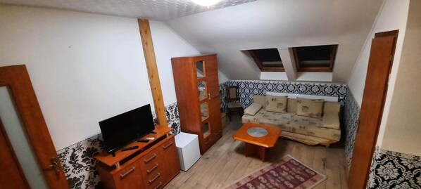 A compact living space with a television on a wooden cabinet, a small white refrigerator, a patterned sofa, and a red rug.