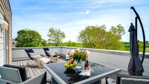 En udendørs terrasse med fletningsmøbler, et glasbord og udsigt til grøn bevoksning.