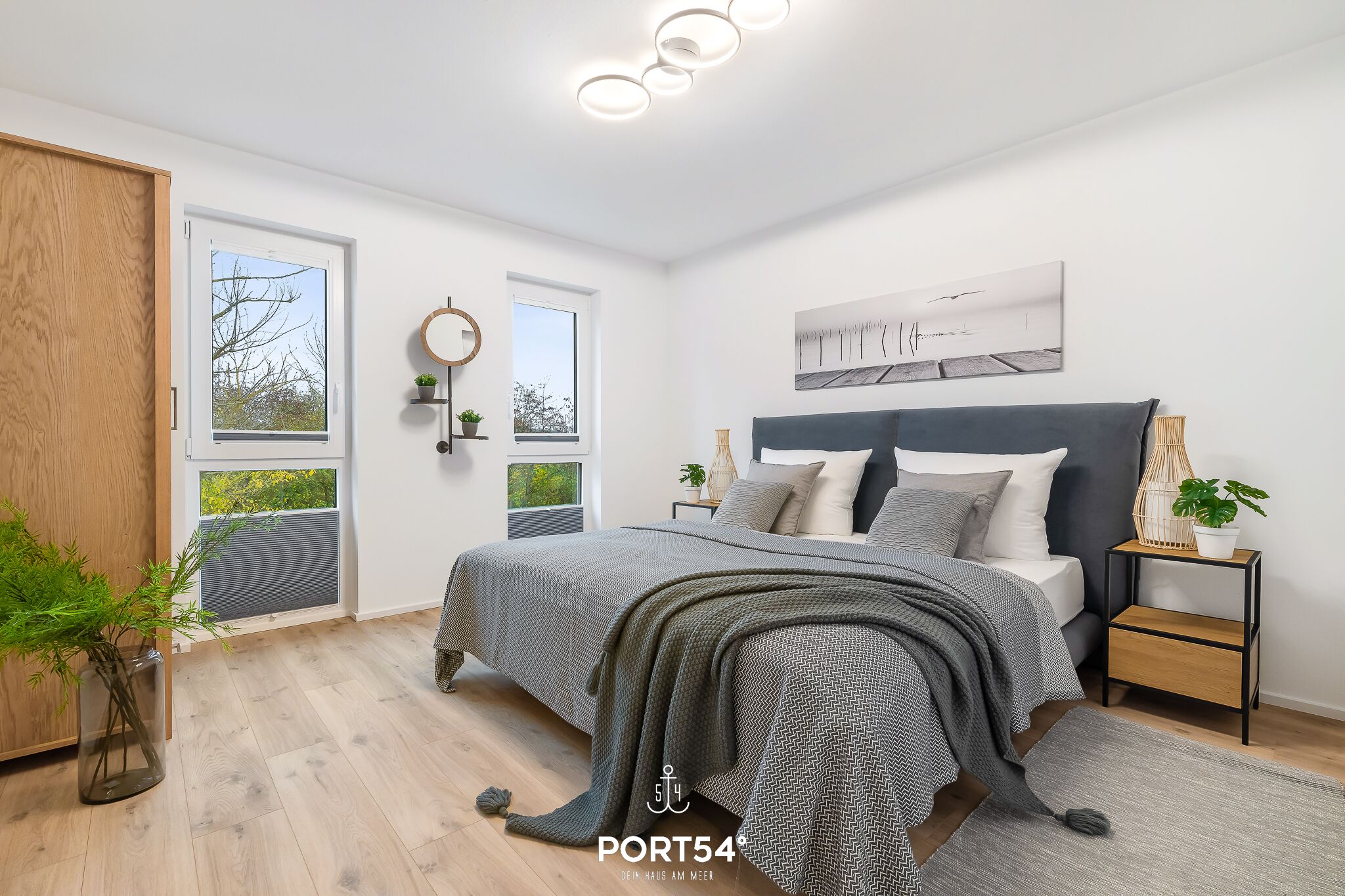 A modern bedroom with a large bed, a bedside table with a plant, a window with a view, and a ceiling light fixture.