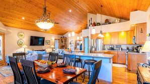 A spacious kitchen with wooden cabinets and a central island, a dining area with blue chairs, and a living room with a sofa and TV.