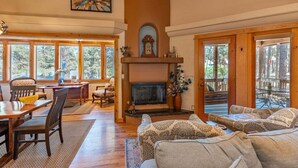 A cozy living room with a fireplace, wooden furniture, and large windows offering a view of the outdoors.