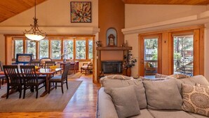 A spacious living room with a high ceiling, wooden floors, a fireplace, and large windows offering a view of the outdoors.