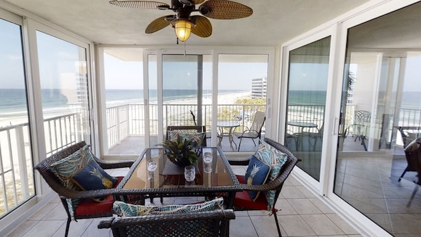 A balcony with a glass door leading to a beach view, wicker furniture, and a ceiling fan.