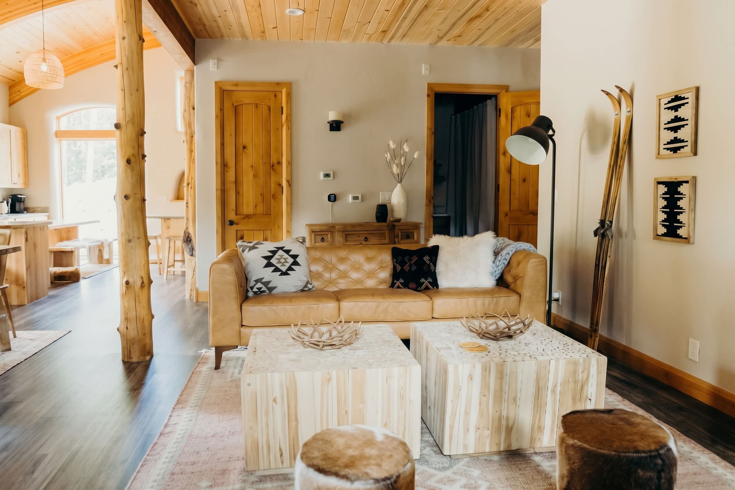 A cozy living room with a sofa, two side tables, and a coffee table. There is a wooden ceiling and a large window.