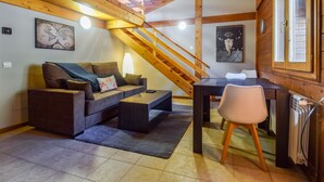 Un salon chaleureux avec un canapé, une table basse et une table à manger avec une chaise. Un escalier en bois mène à l’étage supérieur.