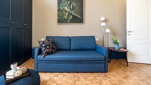 A modern living room with a blue sofa, a decorative pillow, a framed abstract painting, a small side table with a plant, and a white door.