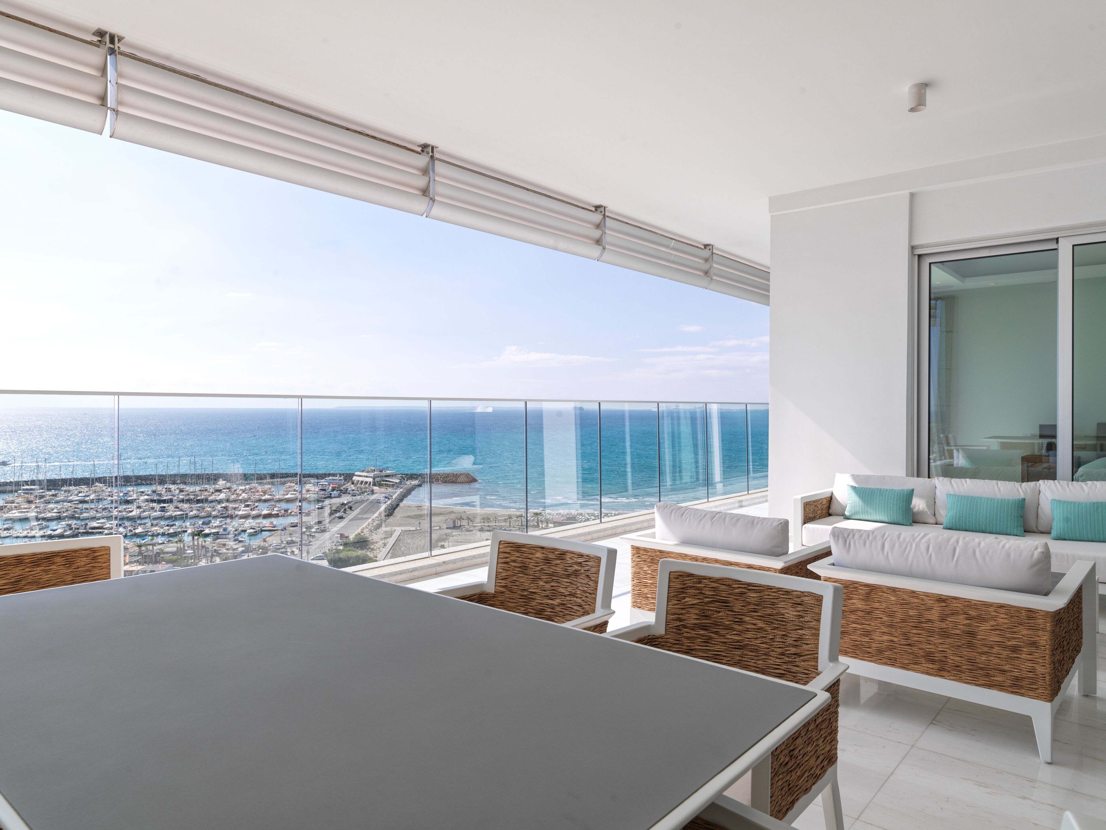 A balcony with a view of the sea and marina. There is a table and chairs with wicker cushions.
