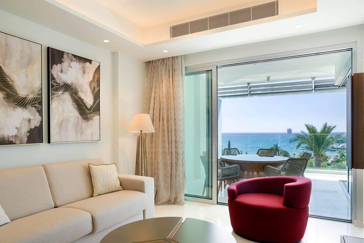 Una sala de estar moderna con un sofá, un sillón rojo, un ventanal de vidrio que da a un balcón con vistas al mar.