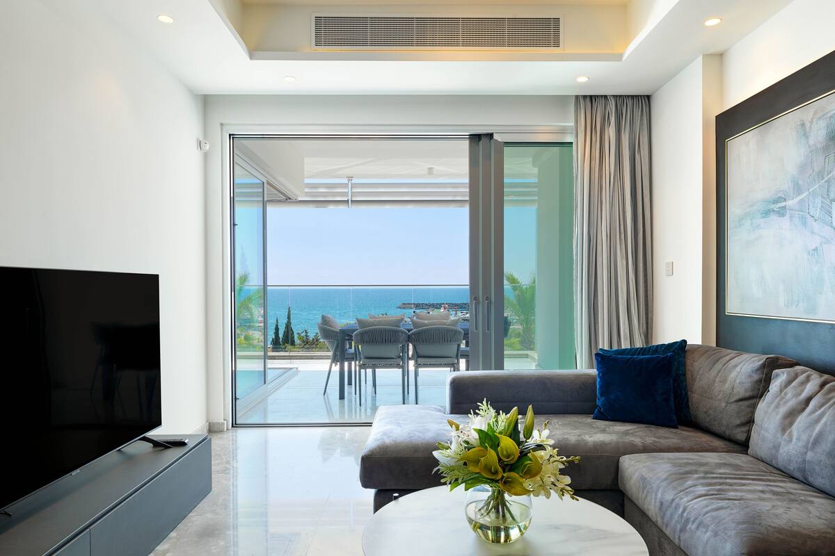 Un salón moderno con una televisión de pantalla plana, un sofá, una mesa de centro con un jarrón de flores y un balcón con vistas al mar.
