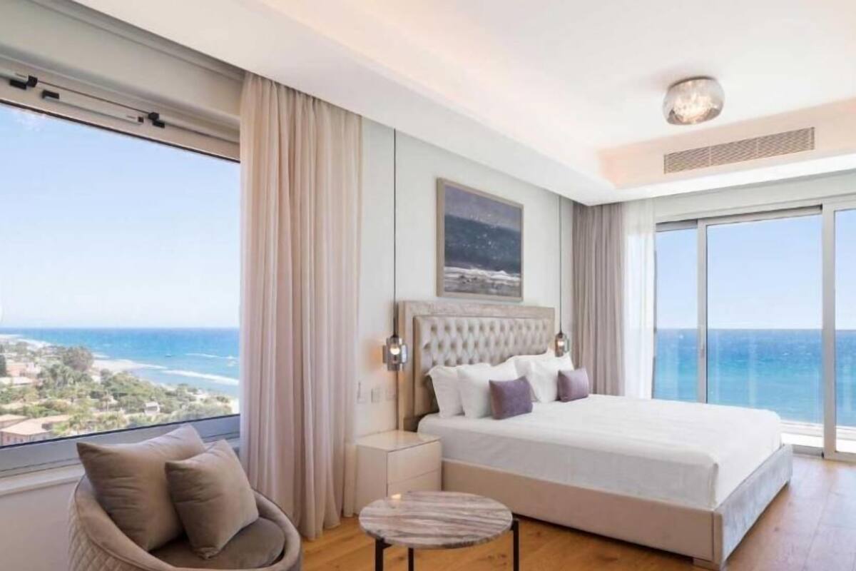 Un dormitorio con una cama grande, vista al océano, una silla y una mesita.