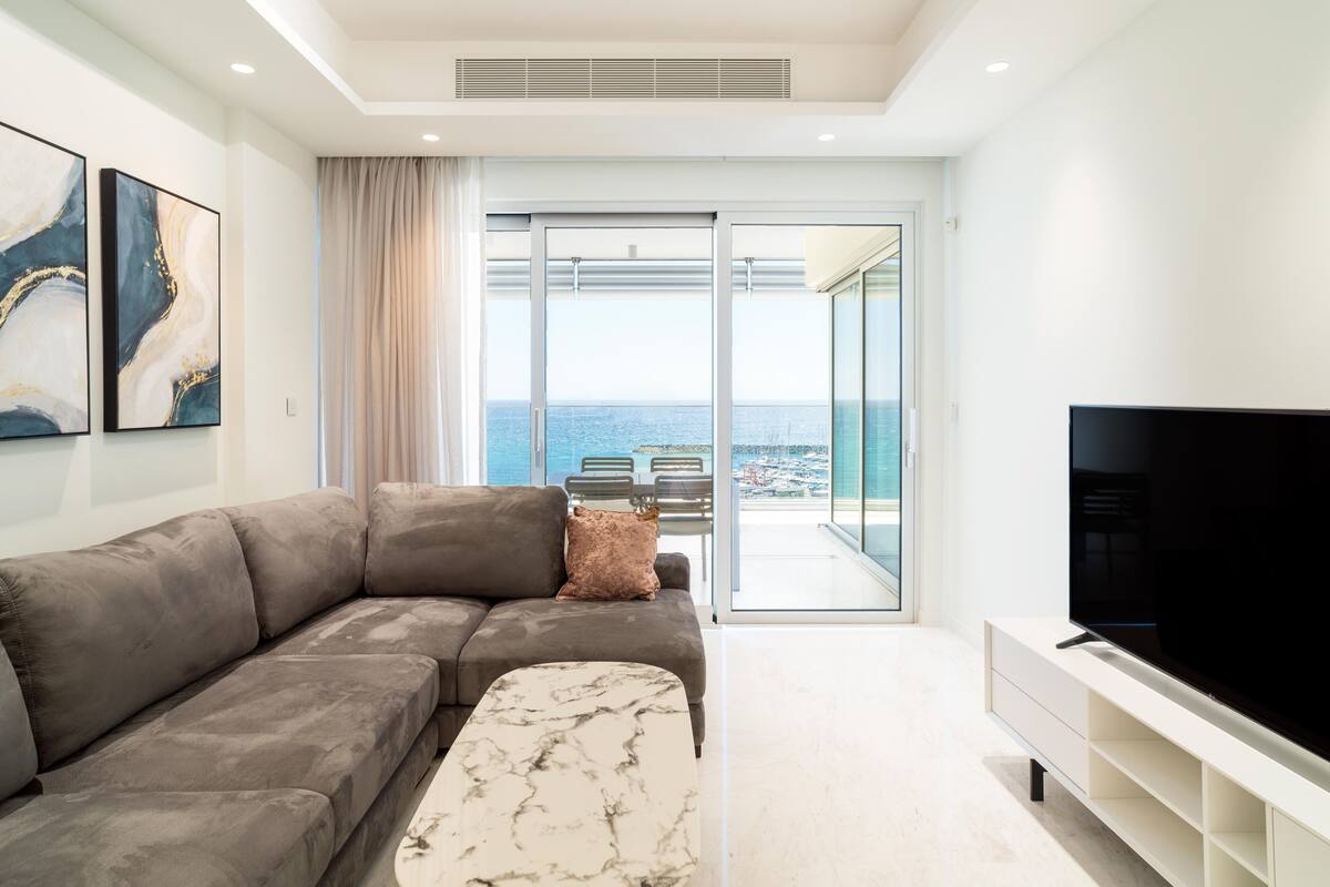 Una sala moderna con un sofá seccional gris, una mesa de centro de mármol, un televisor de pantalla plana sobre un mueble blanco y un balcón con vista al mar.