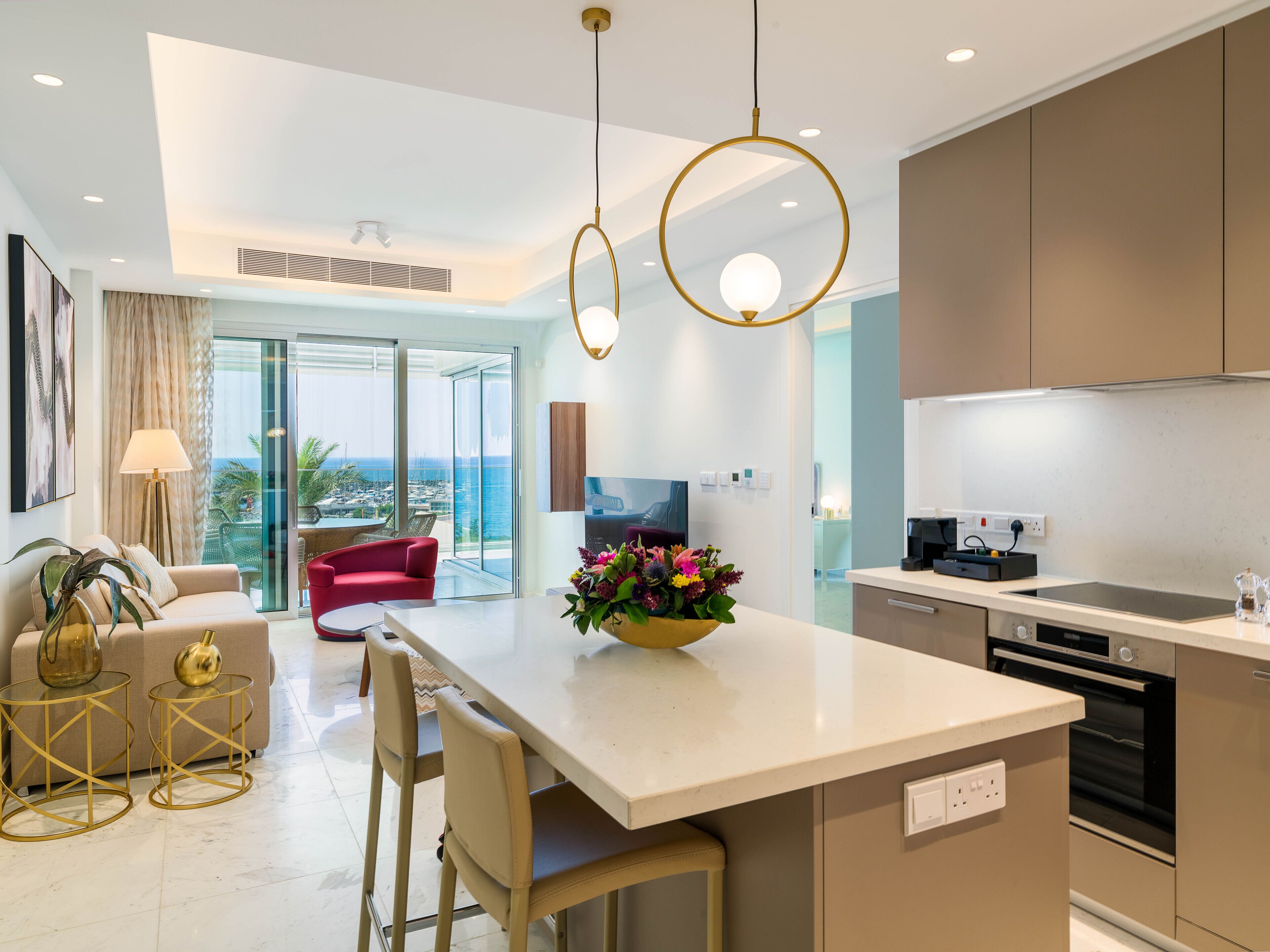 A modern kitchen with a central island, a dining area with a red chair, and a view of the sea through large windows.