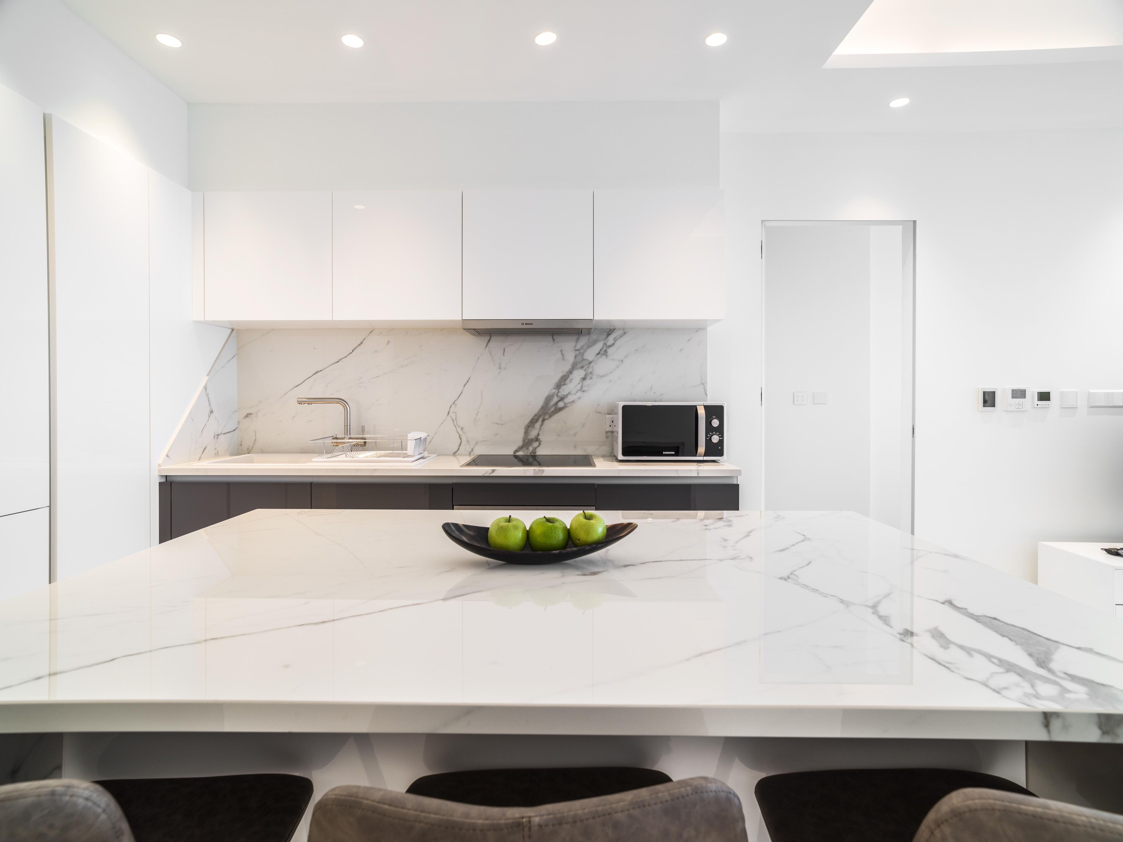 A modern kitchen with a marble countertop, a built-in microwave, and a bowl of green apples on the island.