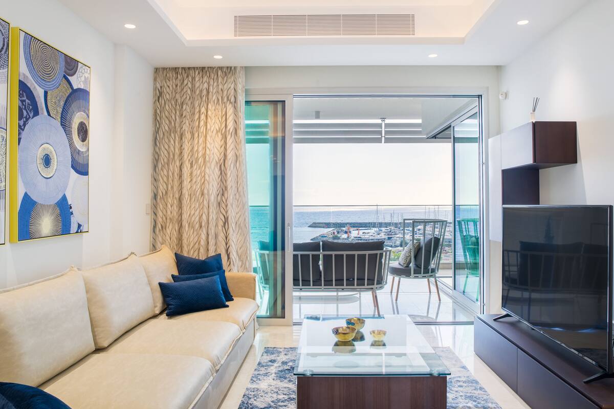 Una sala de estar moderna con sofá, mesa de centro y televisor. Cuenta con un ventanal con vistas al mar y a barcos.