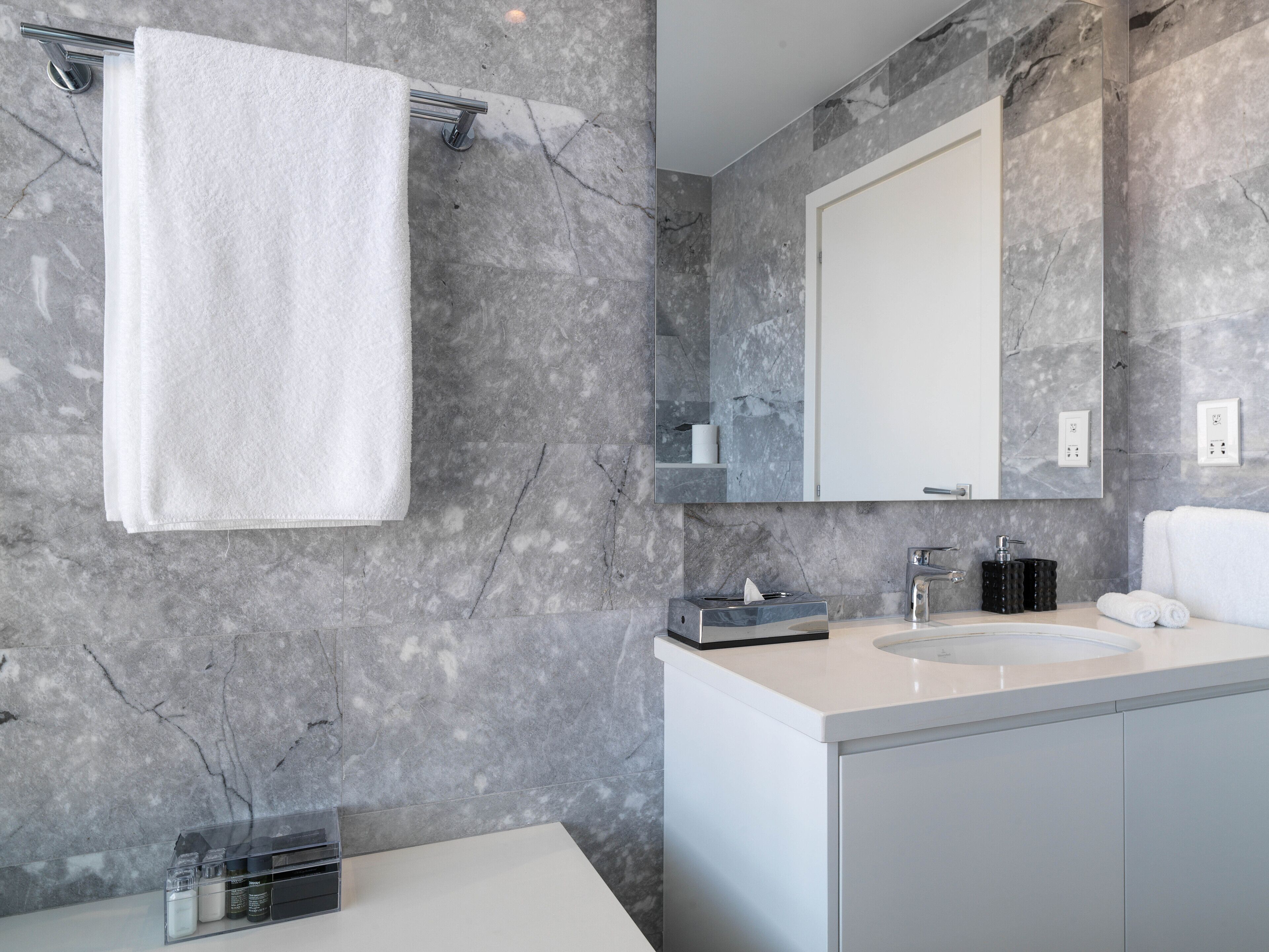 A modern bathroom with a marble wall, a white sink, and a large mirror.