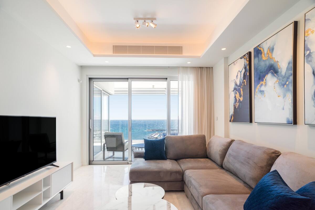 Una sala moderna con un televisor de pantalla plana grande, un sofá seccional color beige, una mesa de centro de mármol y un balcón con vista al mar.