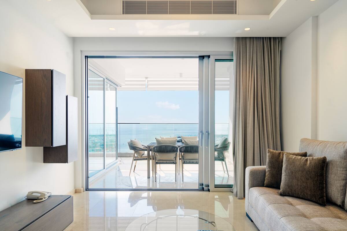 Una sala moderna con una mesa de centro de vidrio, un sofá con cojines, un televisor de pantalla plana en la pared y un balcón con mesa y sillas.