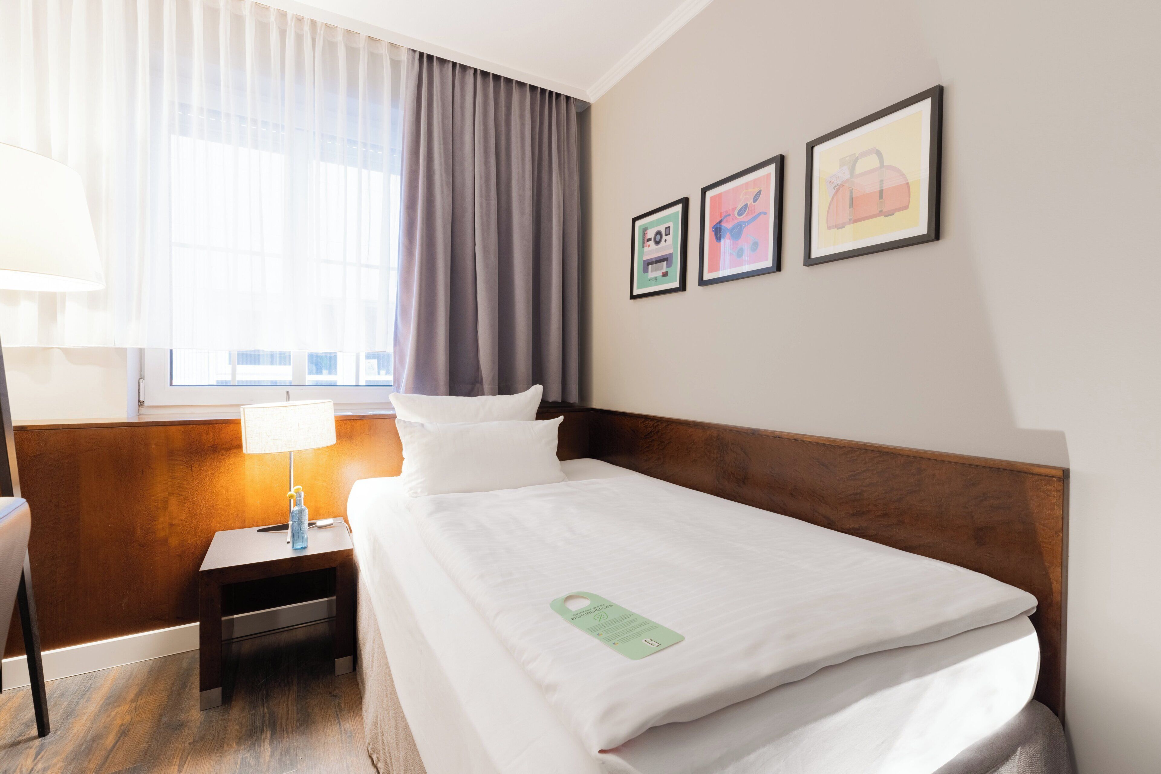 A hotel room with a neatly made bed, a nightstand with a lamp, and two framed pictures on the wall.
