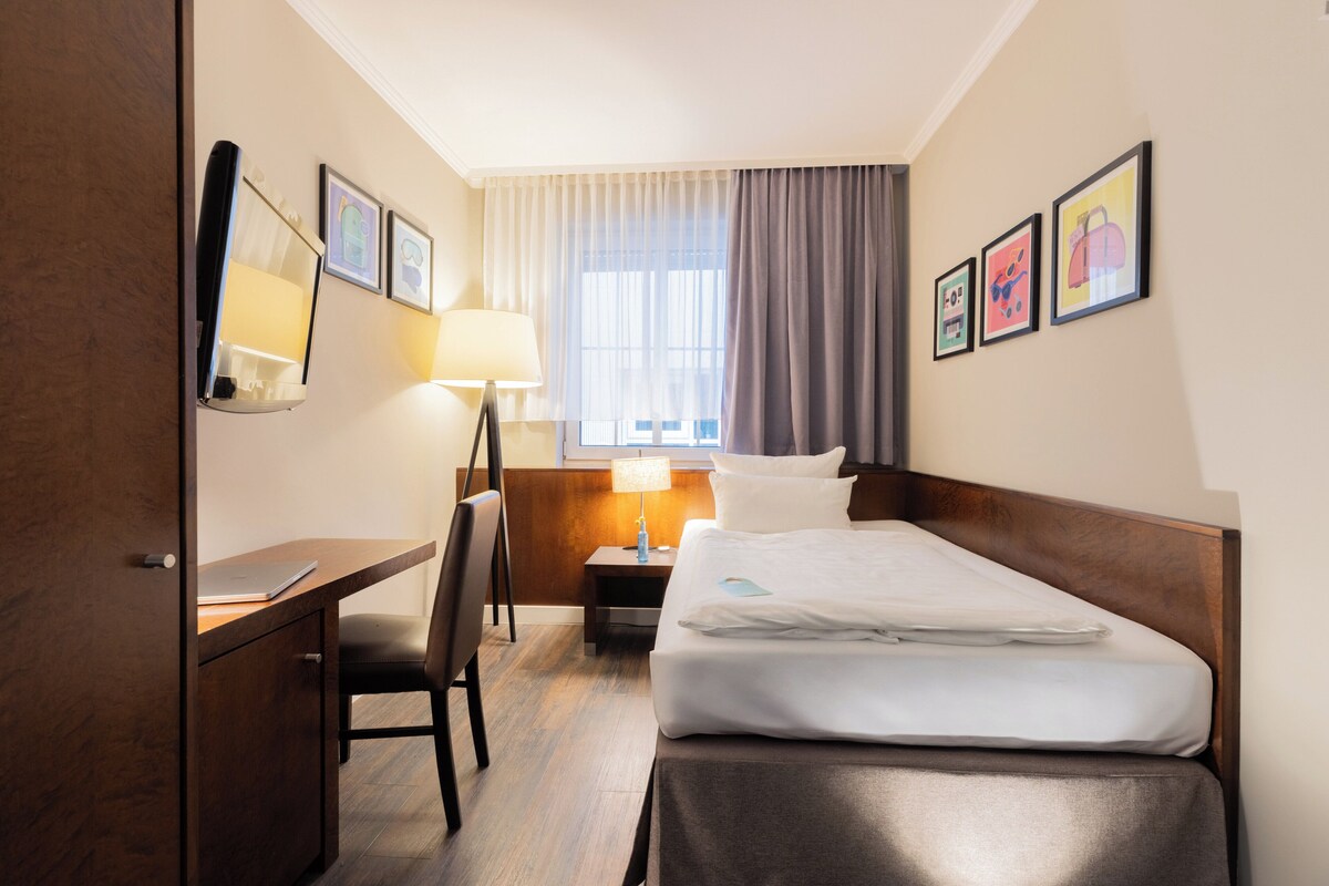 A hotel room with a bed, desk, chair, lamp, and artwork on the walls.