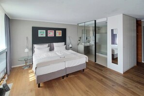 A modern bedroom with a bed, bedside tables, a glass-enclosed walk-in closet, and a large window. - Garner Hotel Friedrichshafen by IHG (Friedrichshafen)
