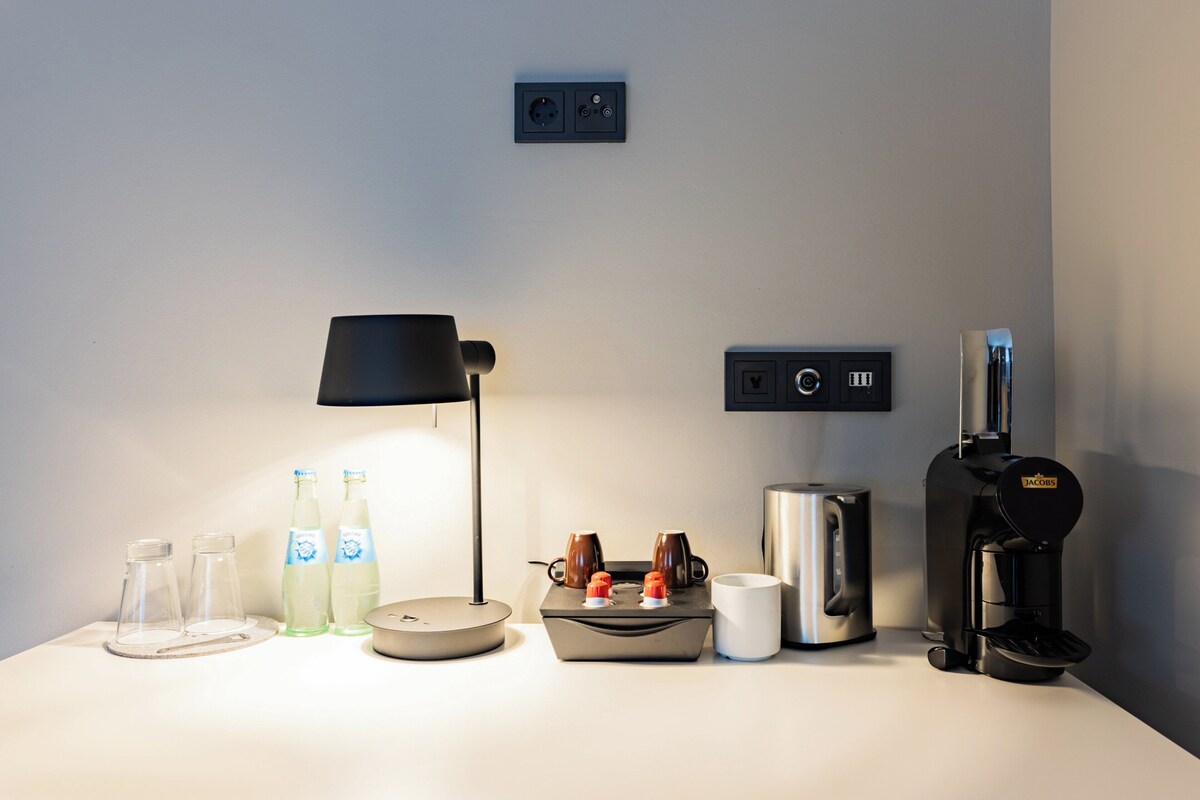 A neatly arranged hotel room with a lamp, two bottles of water, a coffee maker, a kettle, and a small table with cups and a tray.