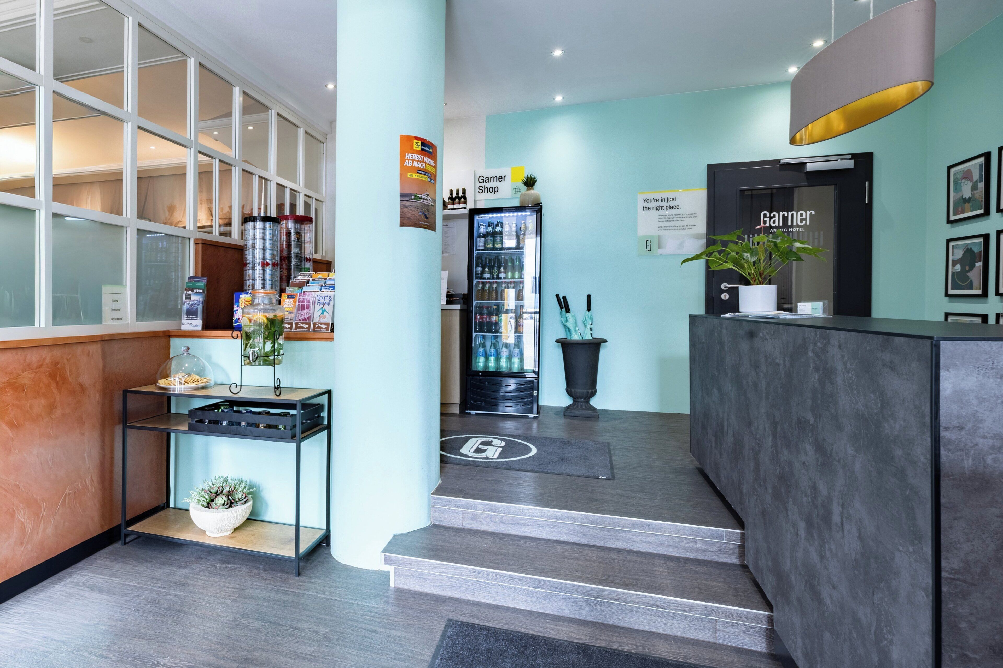 Un espace d’accueil d’hôtel au design moderne, comprenant un comptoir de réception, une vitrine, un réfrigérateur pour boissons et un panneau indiquant l’emplacement de la boutique Garner.
