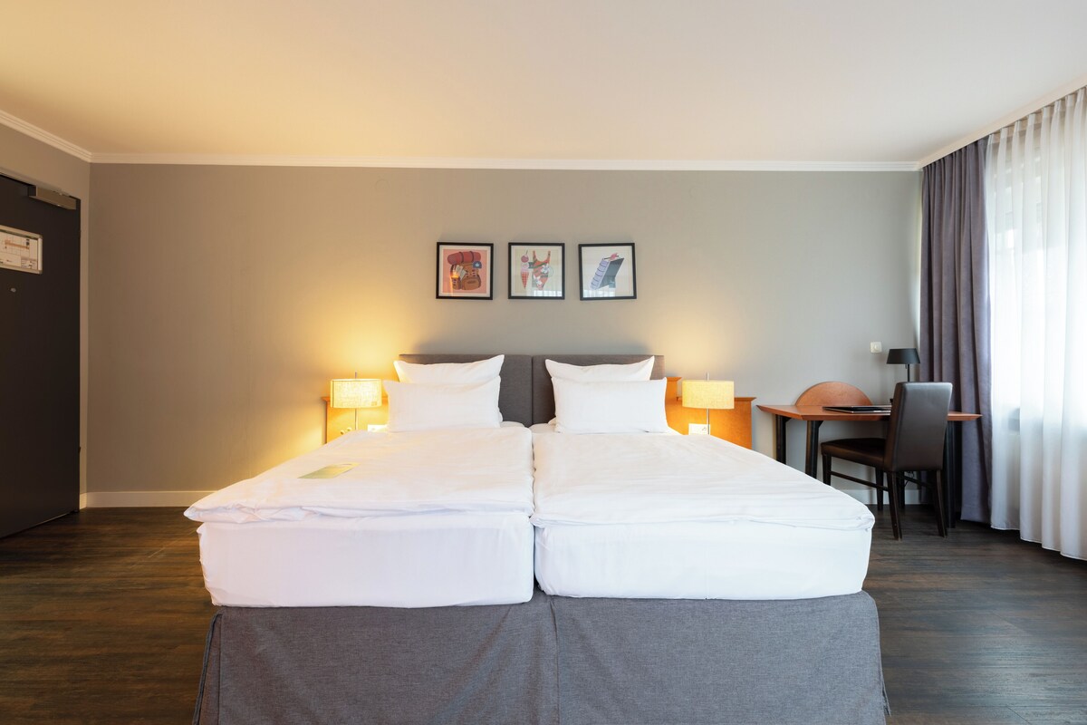 Una cama doble con ropa de cama blanca, un escritorio con una silla y dos lámparas fijadas a la pared.