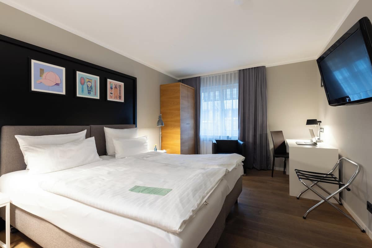 A hotel room with a bed, a desk, a chair, a TV, and artwork on the wall.