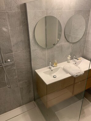 Un baño moderno con ducha de vidrio, un lavabo blanco sobre un mueble de madera y dos espejos redondos.
