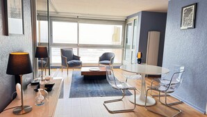 Ein modernes Zimmer mit großem Fenster, einem runden Tisch, zwei Stühlen, einer Lampe und einer Handdesinfektionsflasche.