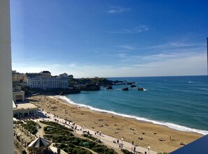 Una playa con un paseo marítimo, un edificio grande y afloramientos rocosos en el mar.