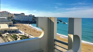 Ein Strand mit freiem Blick auf das Meer, Gebäuden im Hintergrund und einem geschwungenen architektonischen Element im Vordergrund.