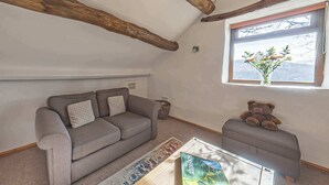 Un salón acogedor con un sofá gris, un pequeño reposapiés y un osito de peluche. Se ve una ventana con vistas a montañas y un jarrón con flores.