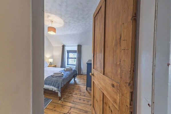 Un dormitorio con puerta de madera, una cama con cubrecama azul, una mesita de noche con lámpara y una ventana con cortinas.