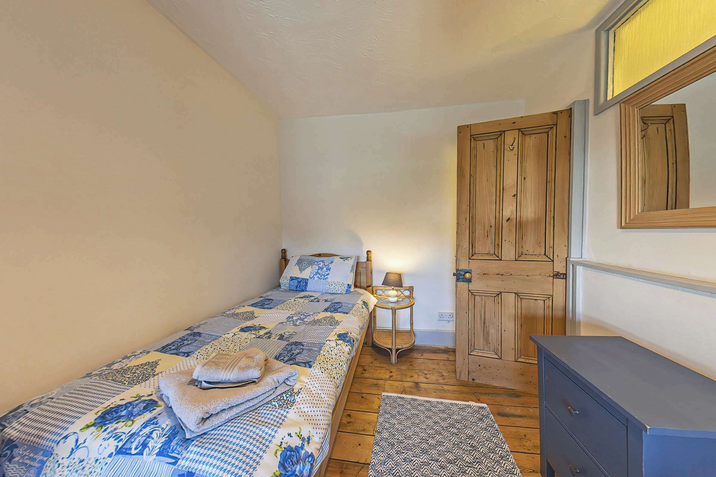 A small, cozy bedroom with a single bed, a bedside table with a lamp, a wooden wardrobe, and a door.