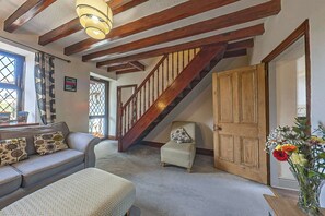 Un salón espacioso con techo alto, vigas de madera a la vista, una escalera, un sofá con cojines estampados y una puerta decorada con arreglos florales.