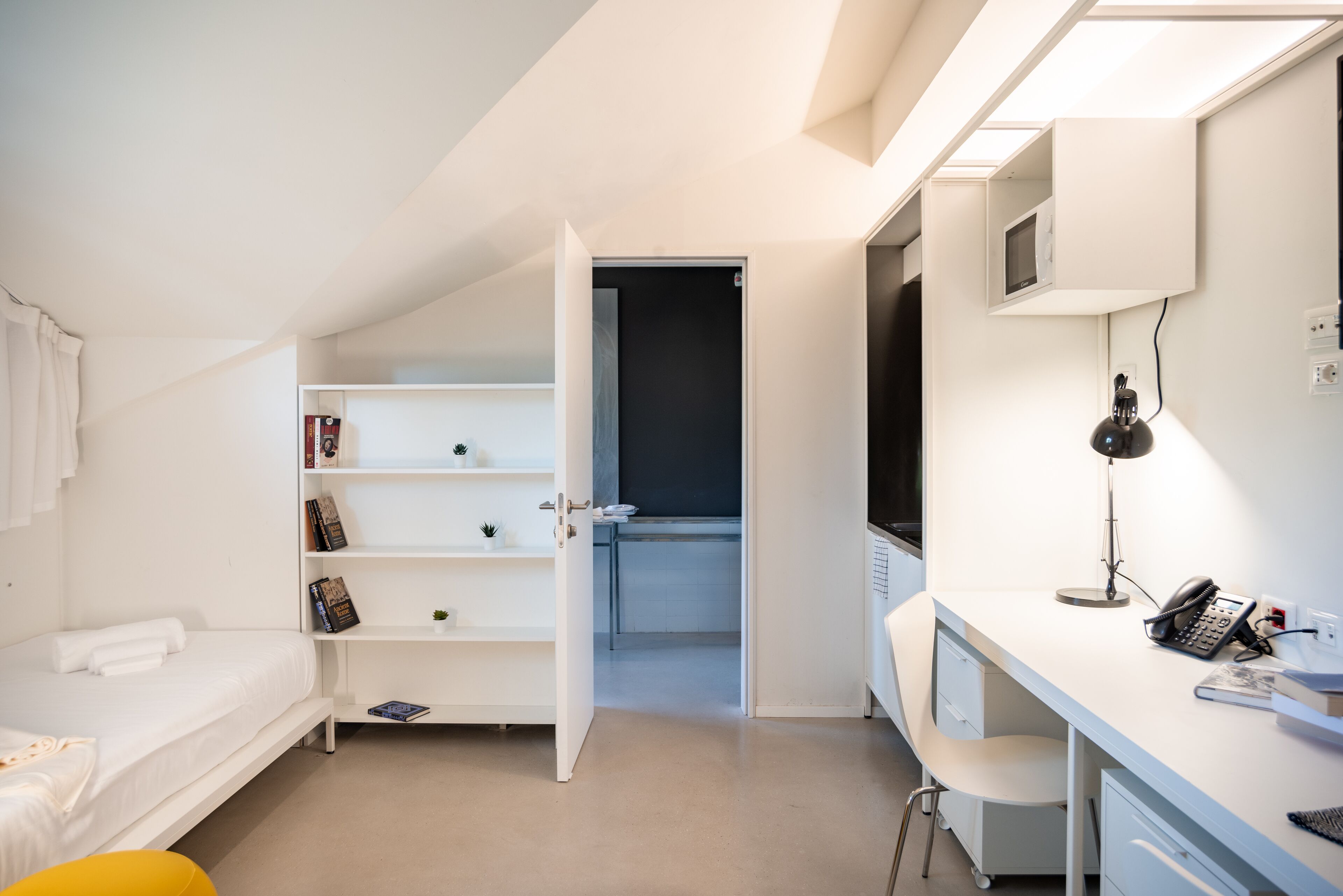 Une chambre moderne avec un lit, un bureau et une chaise. On y trouve également une bibliothèque et une porte qui donne sur une autre pièce.