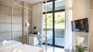 Una habitación de hotel moderna con un amplio ventanal corredero a un balcón, un escritorio con silla, una cama con toallas enrolladas y una planta en maceta.