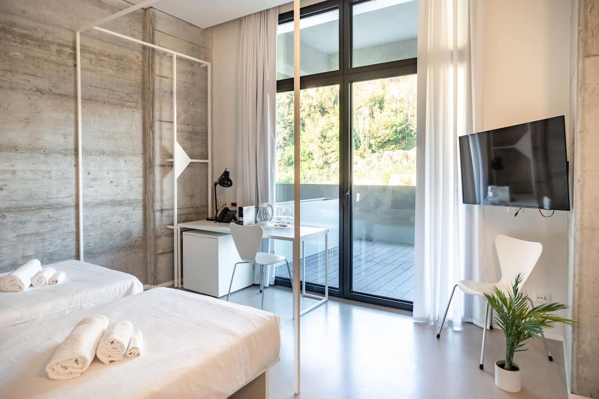 A modern hotel room with a large sliding glass door to a balcony, a desk with a chair, a bed with rolled towels, and a potted plant.