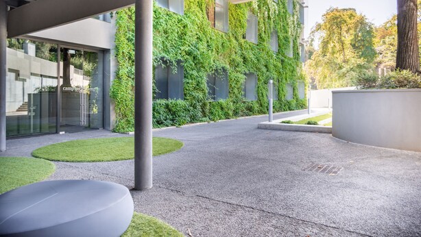 Un edificio moderno con un área exterior techada, suelo de concreto, zona de estar con un banco circular y una pared cubierta de hiedra verde.
