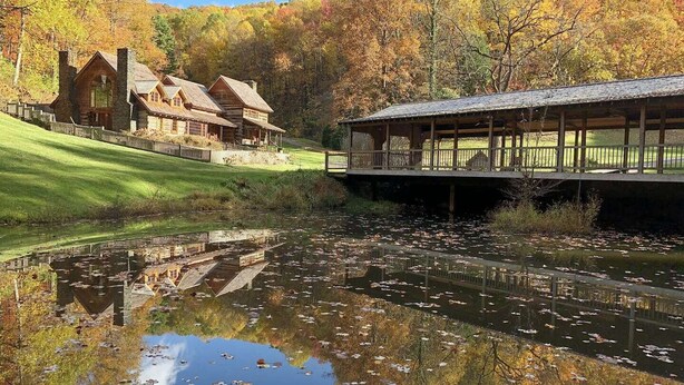 Un chalet rustique doté d’une cheminée en pierre, d’un pont couvert et d’un étang qui reflète le feuillage automnal environnant.