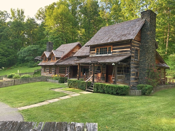 A log cabin with a stone chimney, a porch with wooden chairs, and a stone pathway leading to the entrance.