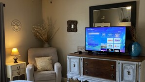 Habitación de hotel con cama, televisión sobre un mueble de madera, lámpara, silla y espejo decorativo.